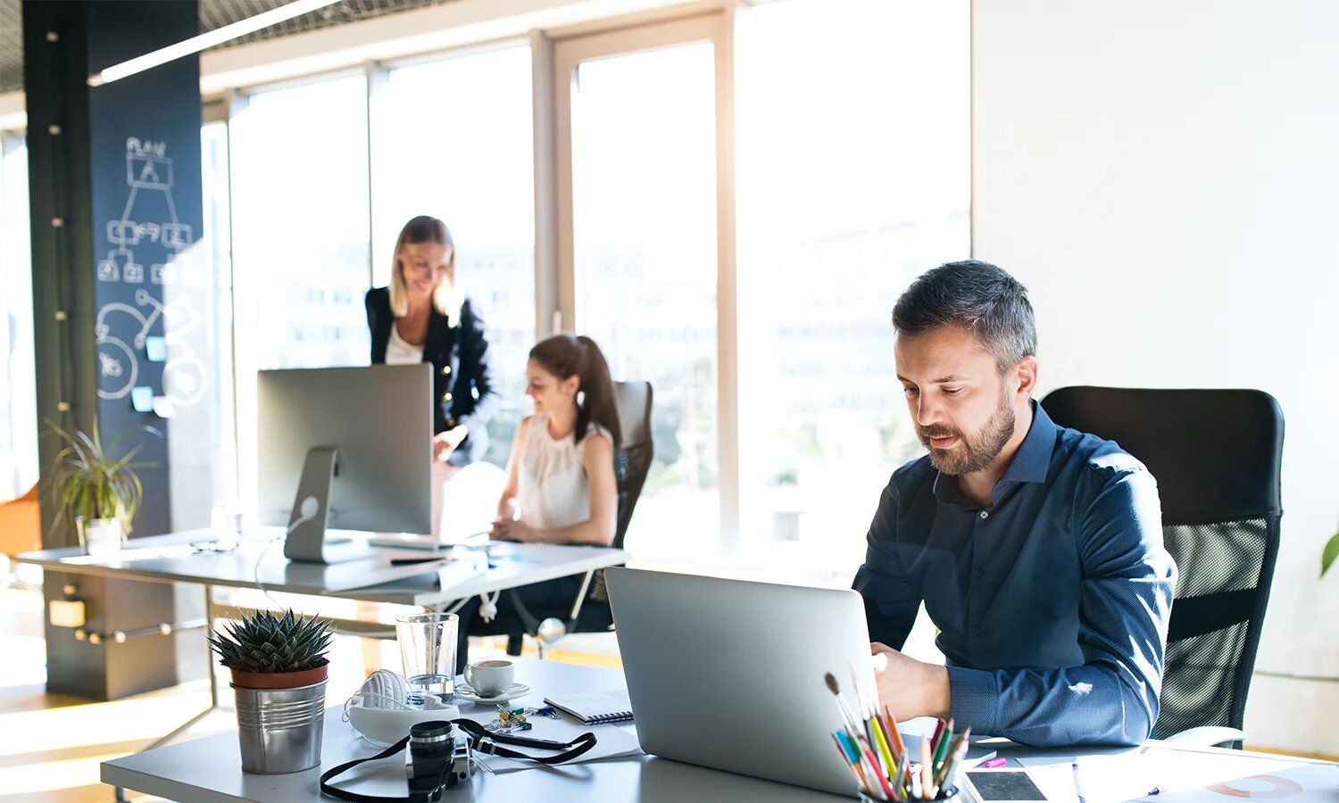 homme qui travaille à son bureau et 2 femmes en arrière plan